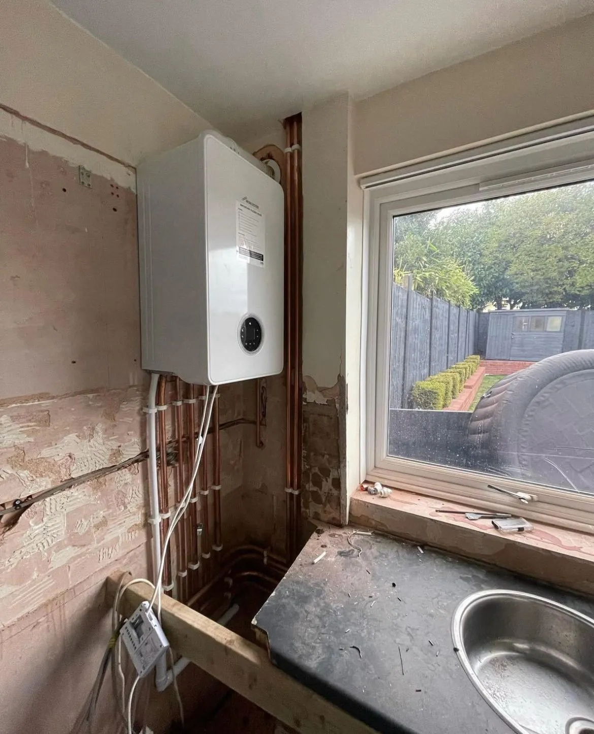 Interior view of a work-in-progress room showing a boiler mounted on the wall, exposed pipes, and a black work surface with a sink.