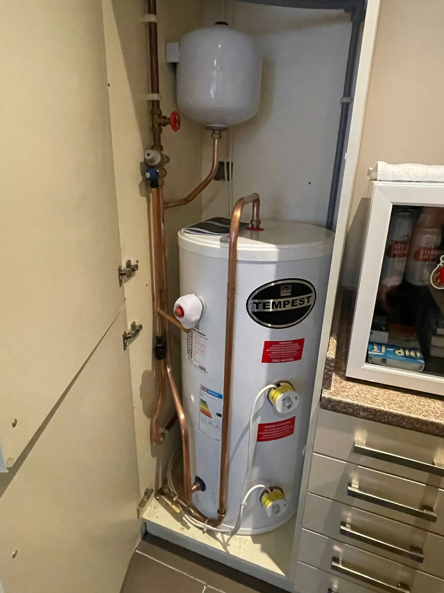 A white water heater with copper piping and a pressure tank, installed in a kitchen cabinet beside drawers and a window.