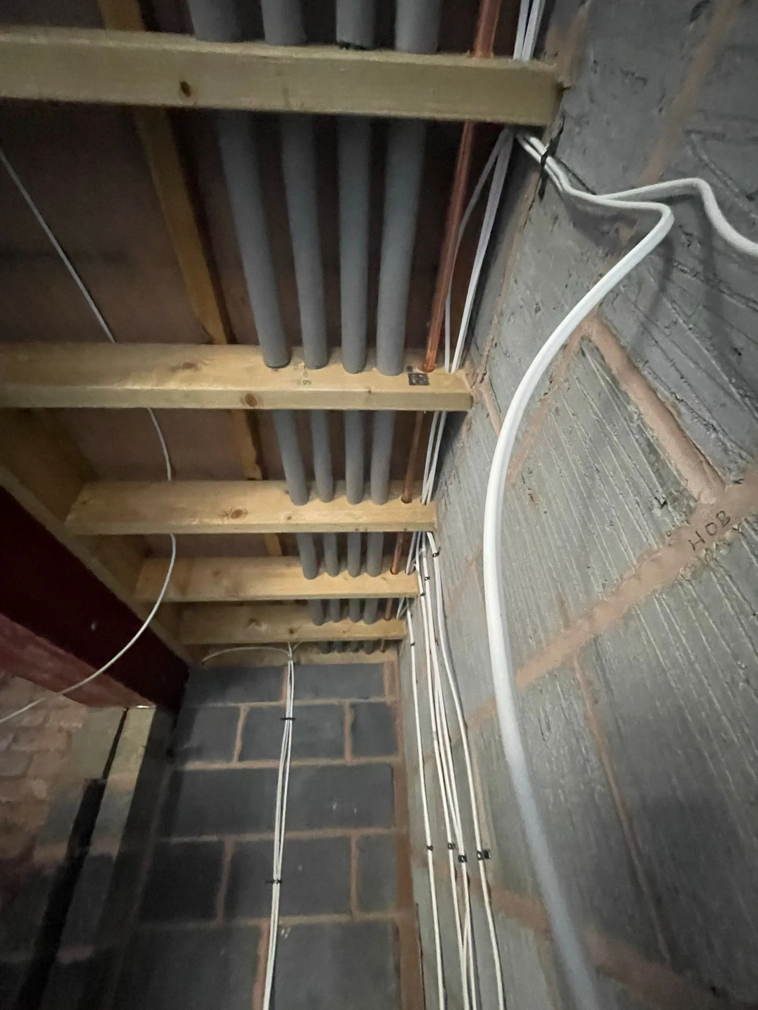 View of a basement ceiling showing wooden beams, gray pipes, and white electrical wires along a concrete wall.