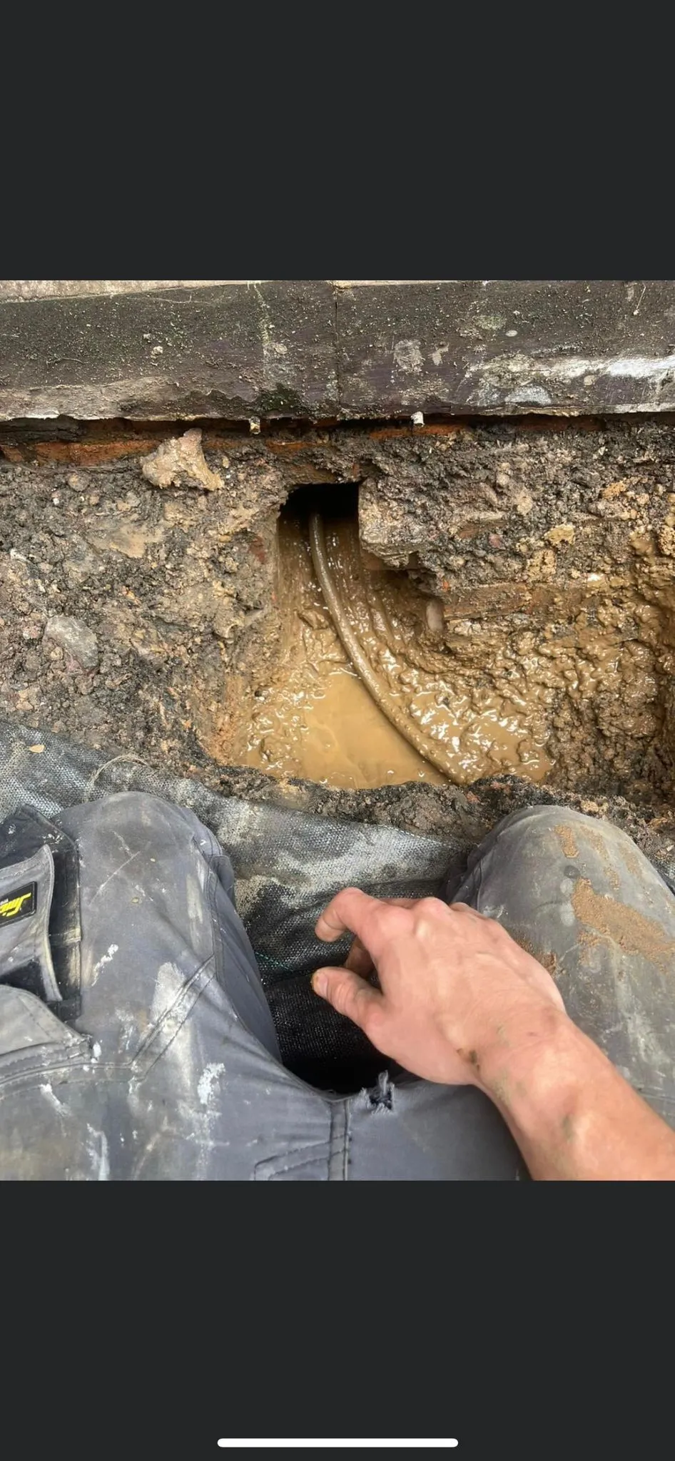A person's legs in work pants are shown near a muddy hole revealing a pipe beneath a foundation.