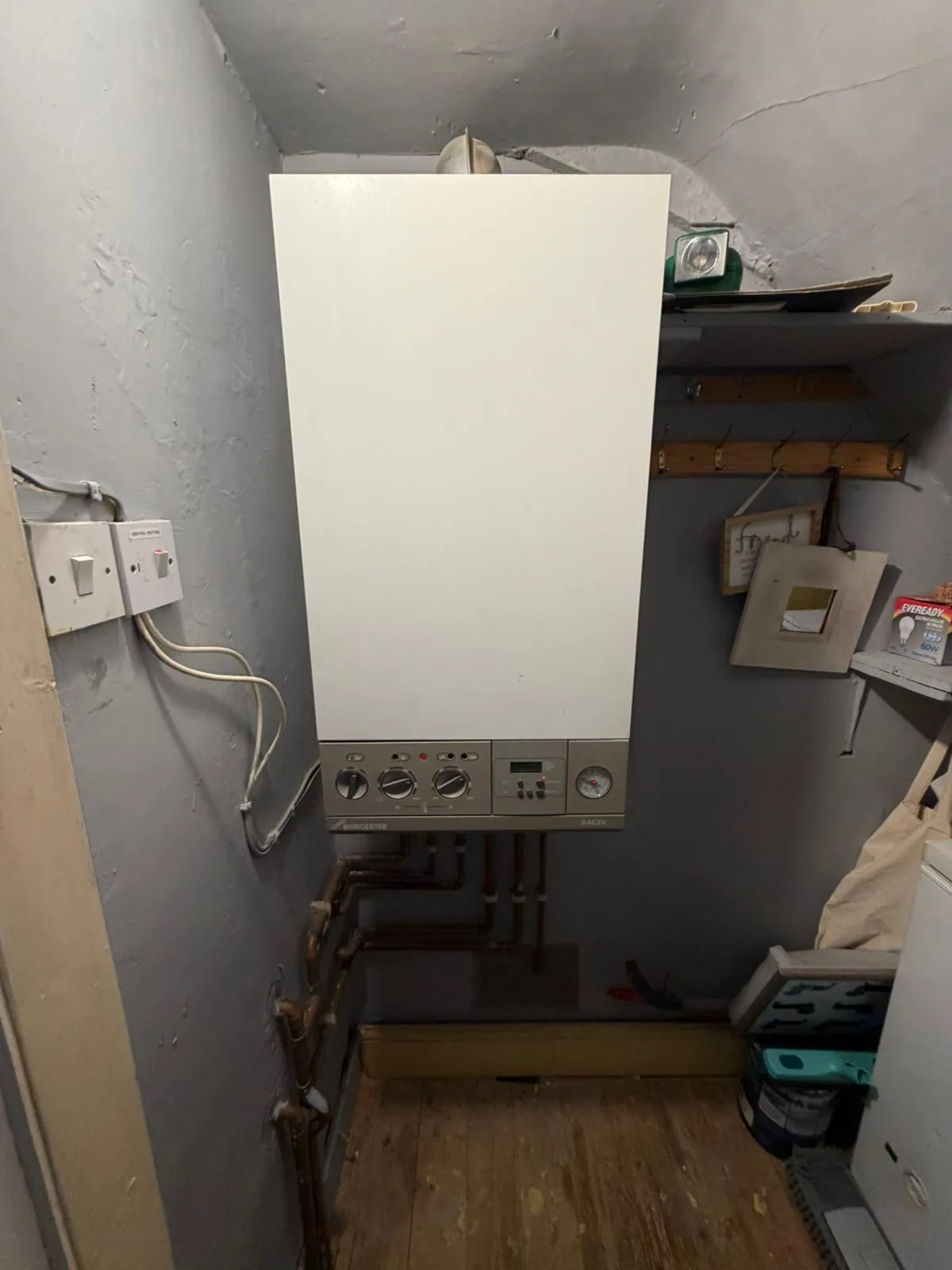 A compact boiler mounted on a wall in a small utility room with exposed pipes and shelves above.