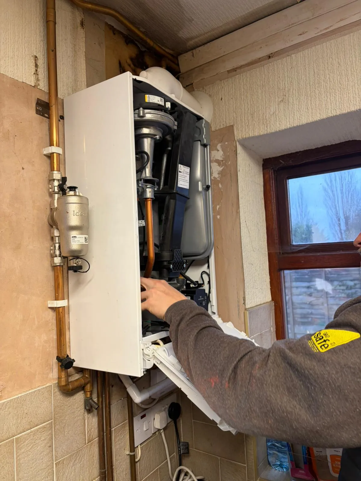 A person is inspecting the inside of a boiler mounted on a wall, with visible pipes and a window in the background.
