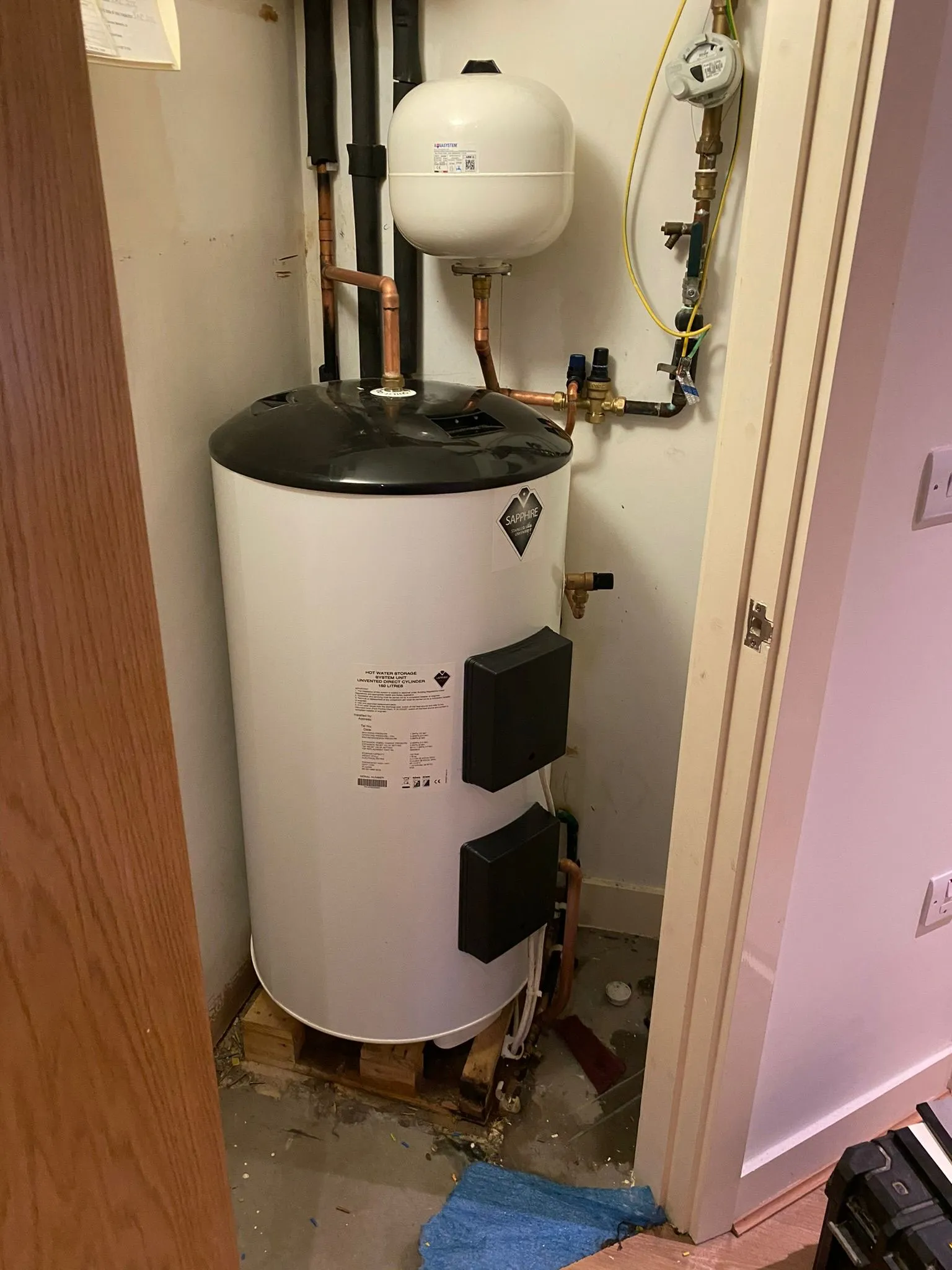 A white hot water tank on wooden pallets, with copper pipes, located in a small utility space with tools and a blue mat nearby.