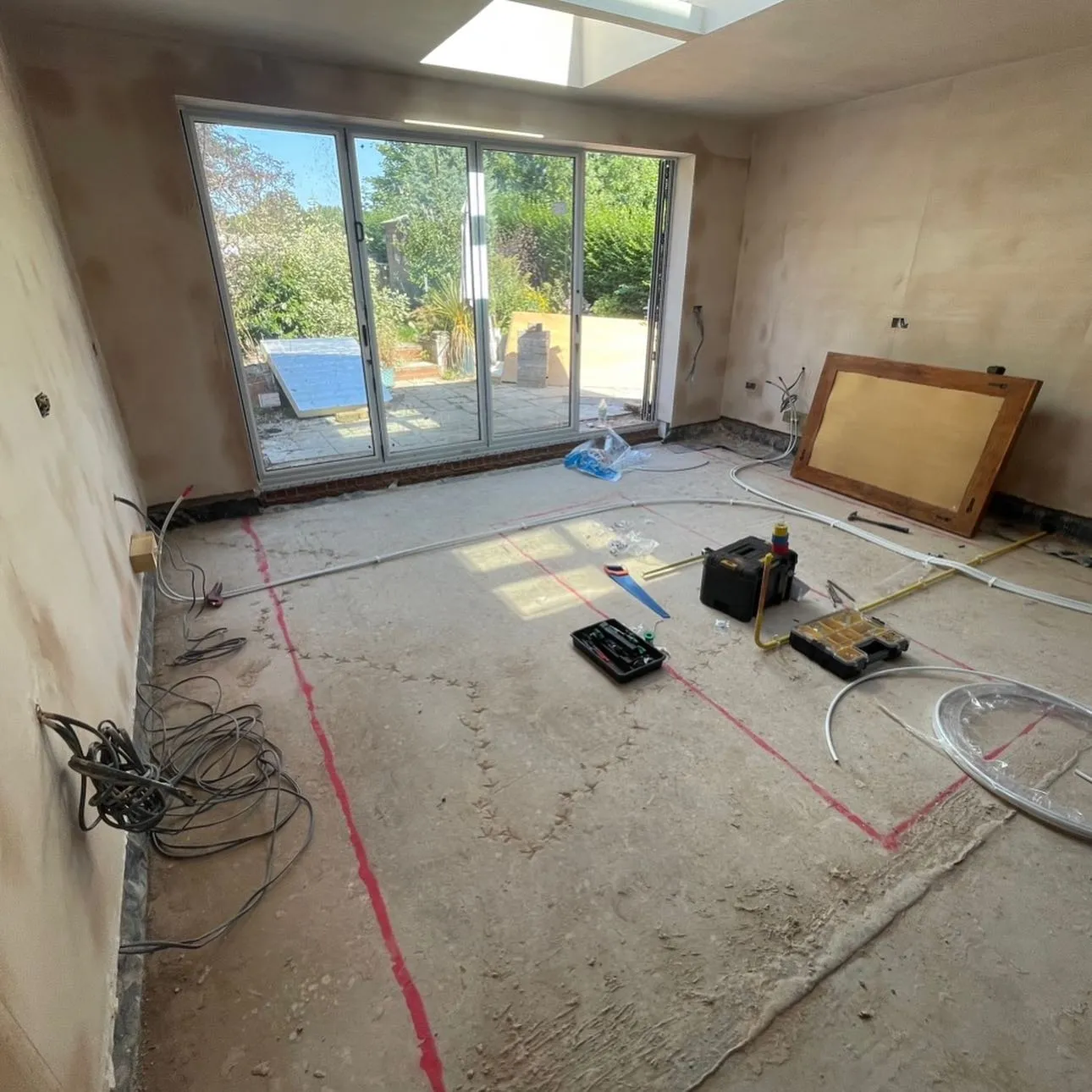 Empty room under renovation with exposed floor, tools scattered around, and sliding glass doors leading to a garden view outside.