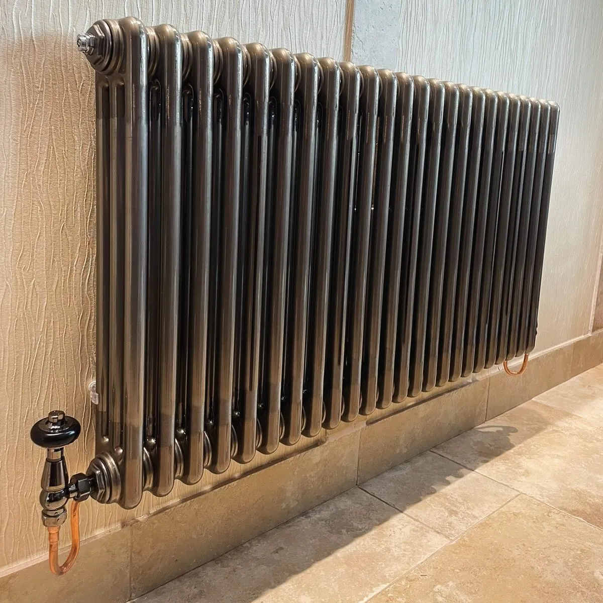 A sleek, modern radiator with multiple vertical panels, installed against a textured wall, featuring a black valve and copper piping.