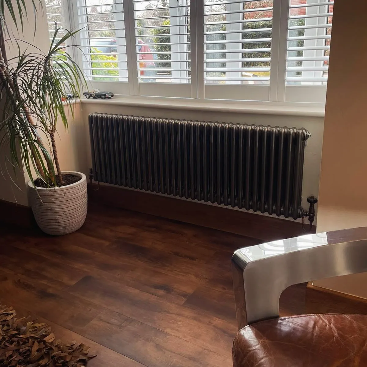A cozy interior corner featuring a potted plant, a vintage radiator, and a stylish chair on wooden flooring, with light coming through the window.