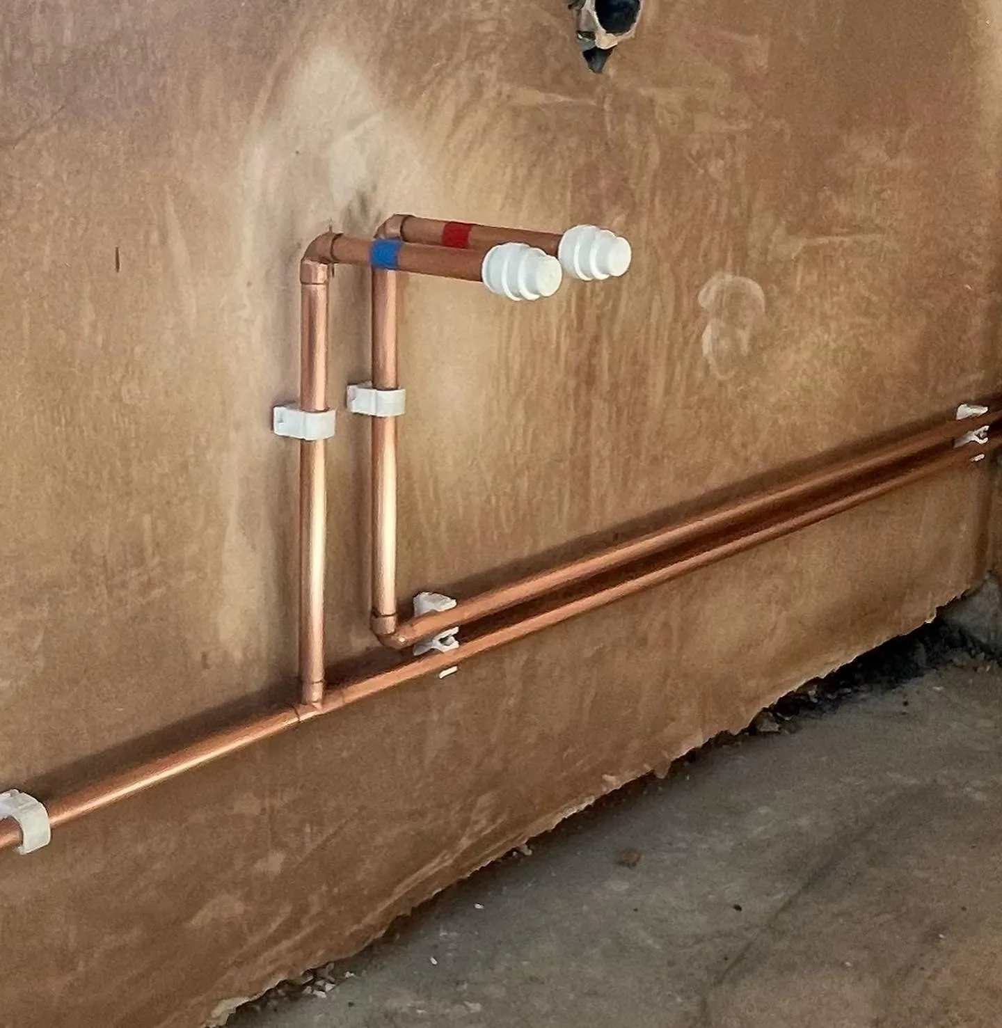 Copper plumbing pipes are installed against a wall, with two vertical pipes at the top and horizontal pipes below, secured with brackets.
