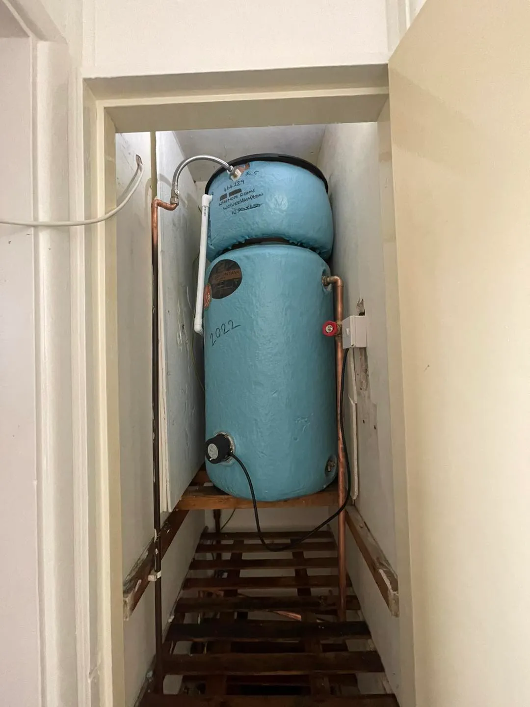 A narrow space showing two stacked blue water tanks with pipes and electrical connections, positioned on a wooden platform.